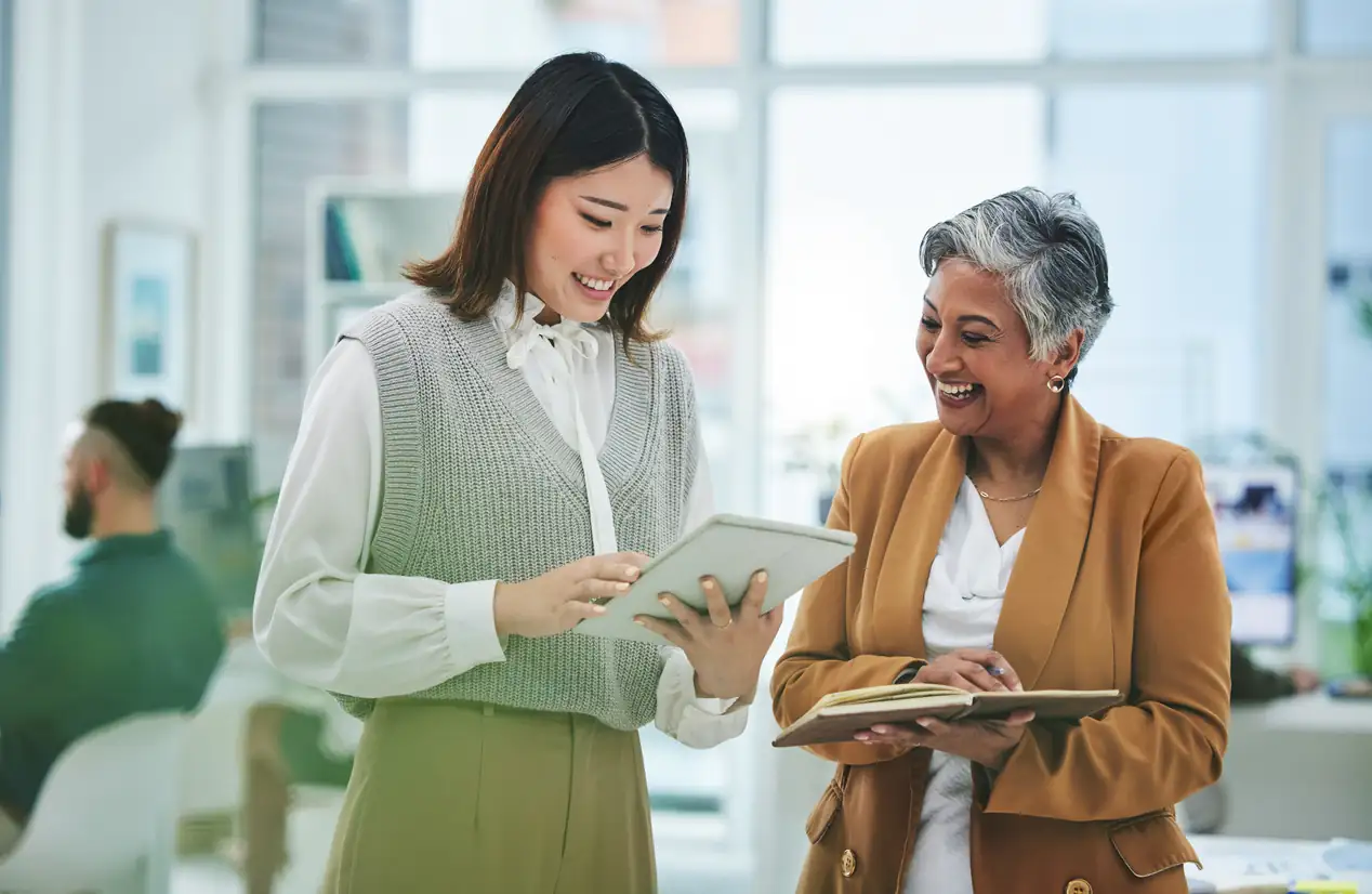 Two women colleagues smiling as they review a tablet and notebook together at work